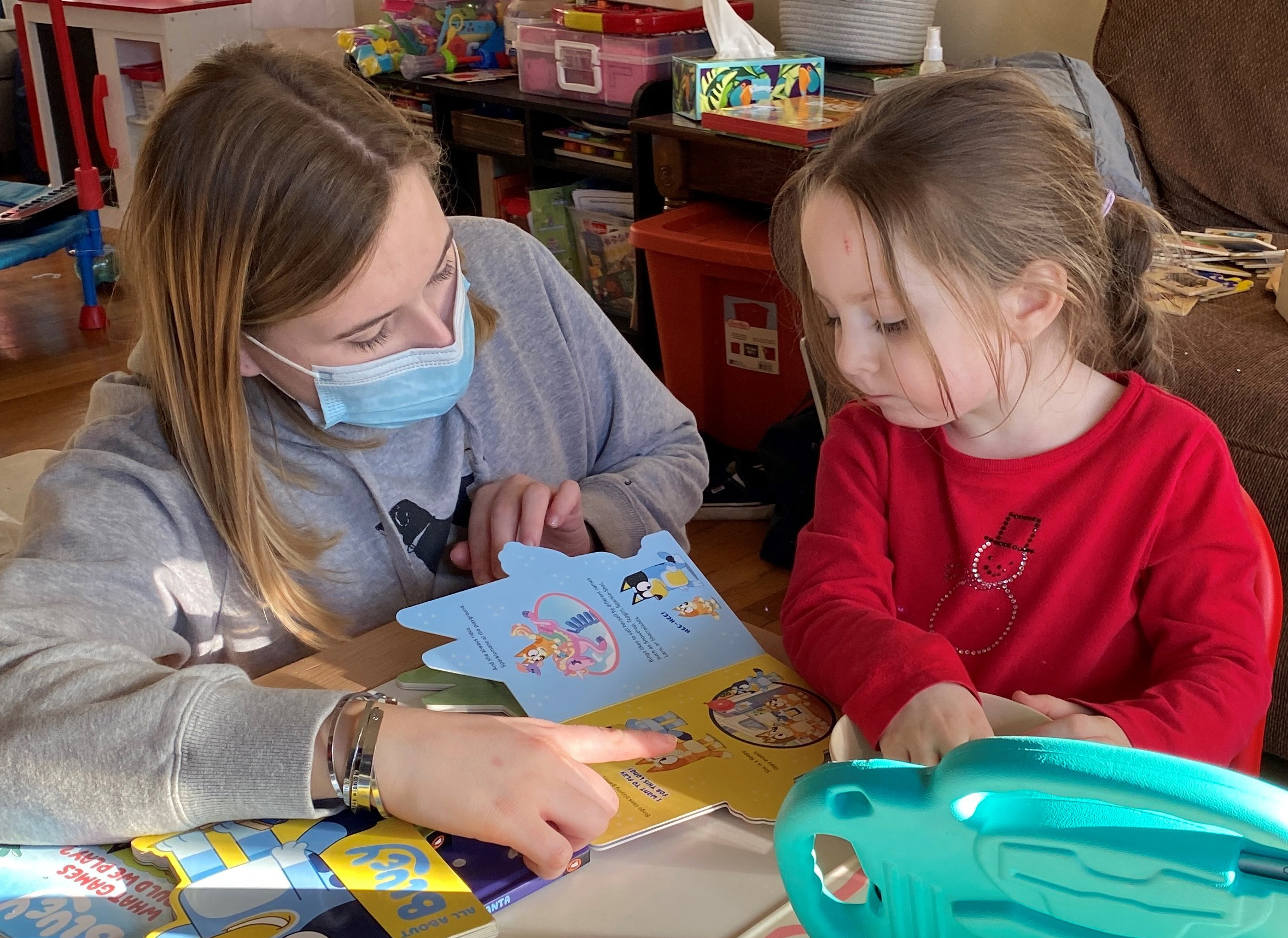 therapist wearing a mask reads to little girl with red shirt