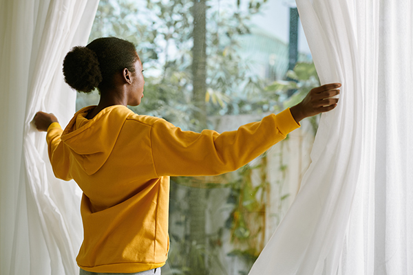 woman in orange sweatshirt opens white curtains to sunny day